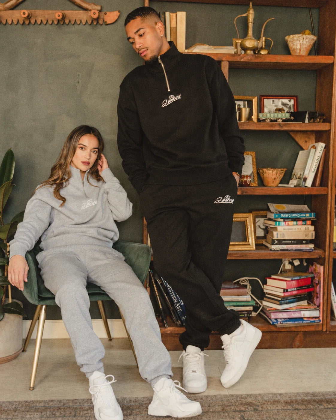 Two models wearing gray and black Hoopery tracksuits beside a bookcase